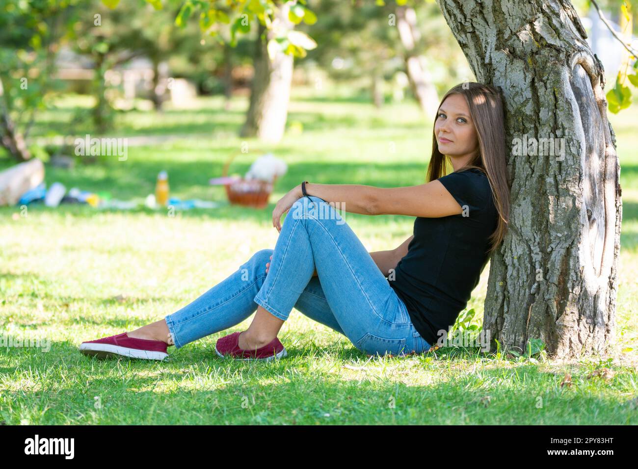 Ein Mädchen sitzt unter einem Baum in einem sonnigen Park und blickt freudig in den Rahmen Stockfoto