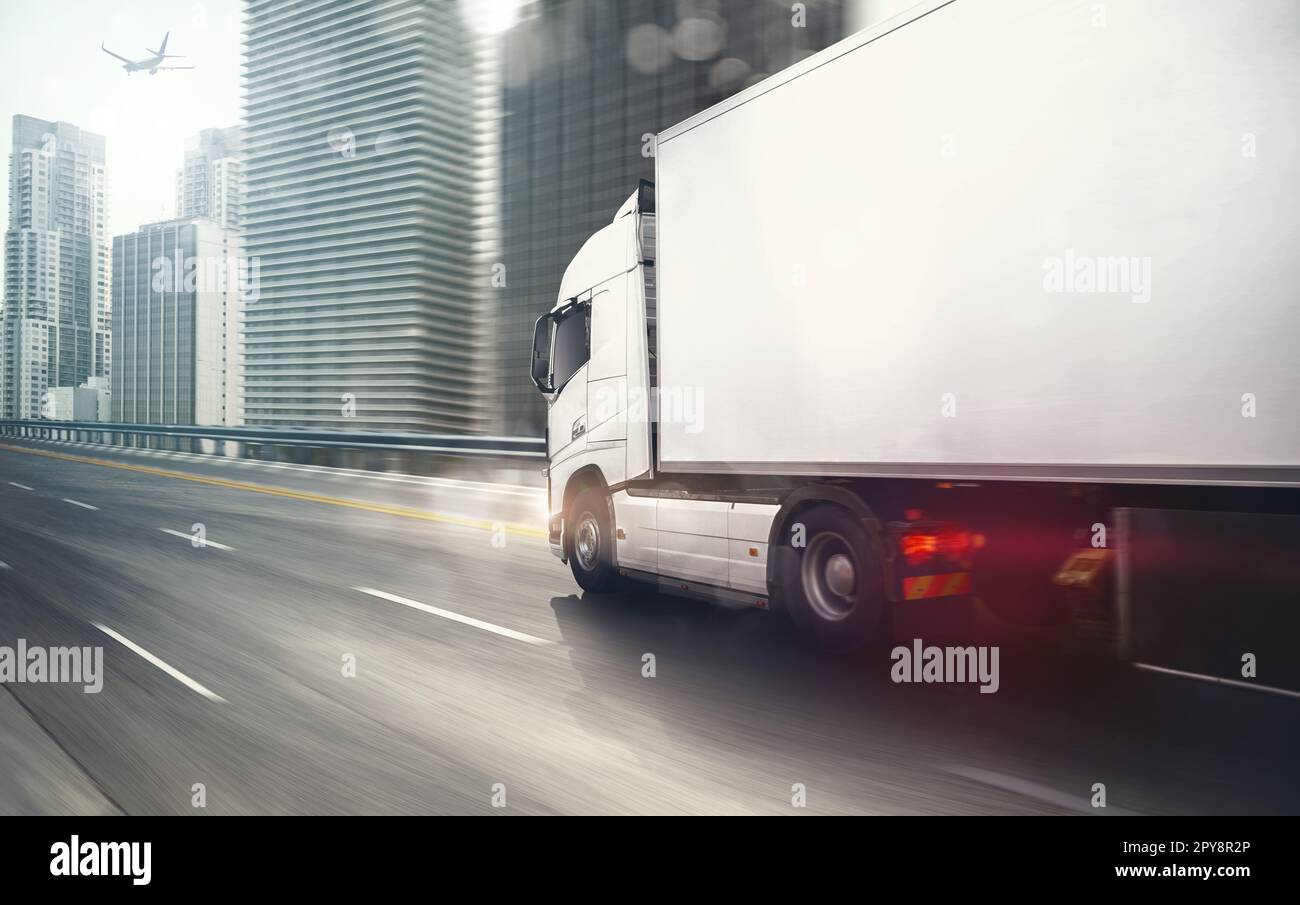 Weißer LKW, der sich schnell auf der Autobahn bewegt, mit einer modernen Stadt im Hintergrund Stockfoto