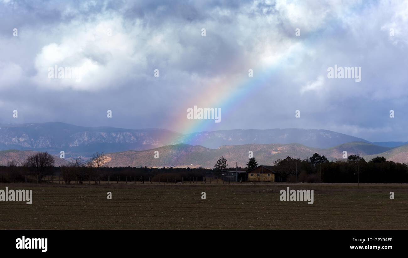 Heller Regenbogen vor dem Hintergrund einer Sturmwolke über der Bergkette und dem Tal. Raue Berglandschaft vor Regen Stockfoto