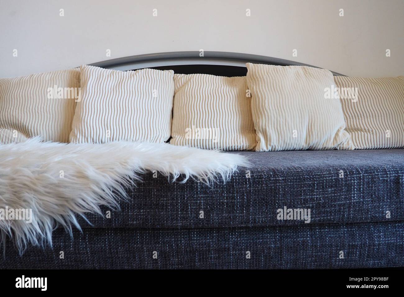 Graues Sofa mit grobem Polstertuch und weißen dekorativen, gewellten Kissen. Über den Sitz wird ein weißer Kunstpelzteppich oder ein Überwurf geschleudert. Wohnzimmereinrichtung Stockfoto