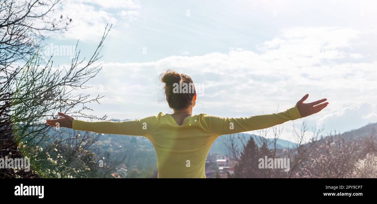 Eine Frau mit ausgestreckten Armen grüßt die Natur, während sie auf dem Hügel auf dem Land steht. Stockfoto