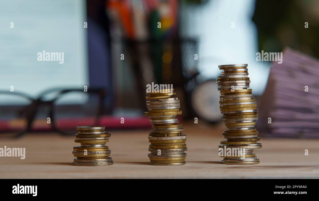 Zusammenstellung von drei Stapeln Münzen neben einem Stapel Papier, einer Brille und einem Laptop auf dem Schreibtisch. Arbeitsplatz, Geschäft, Geld, Bankwesen. Stockfoto