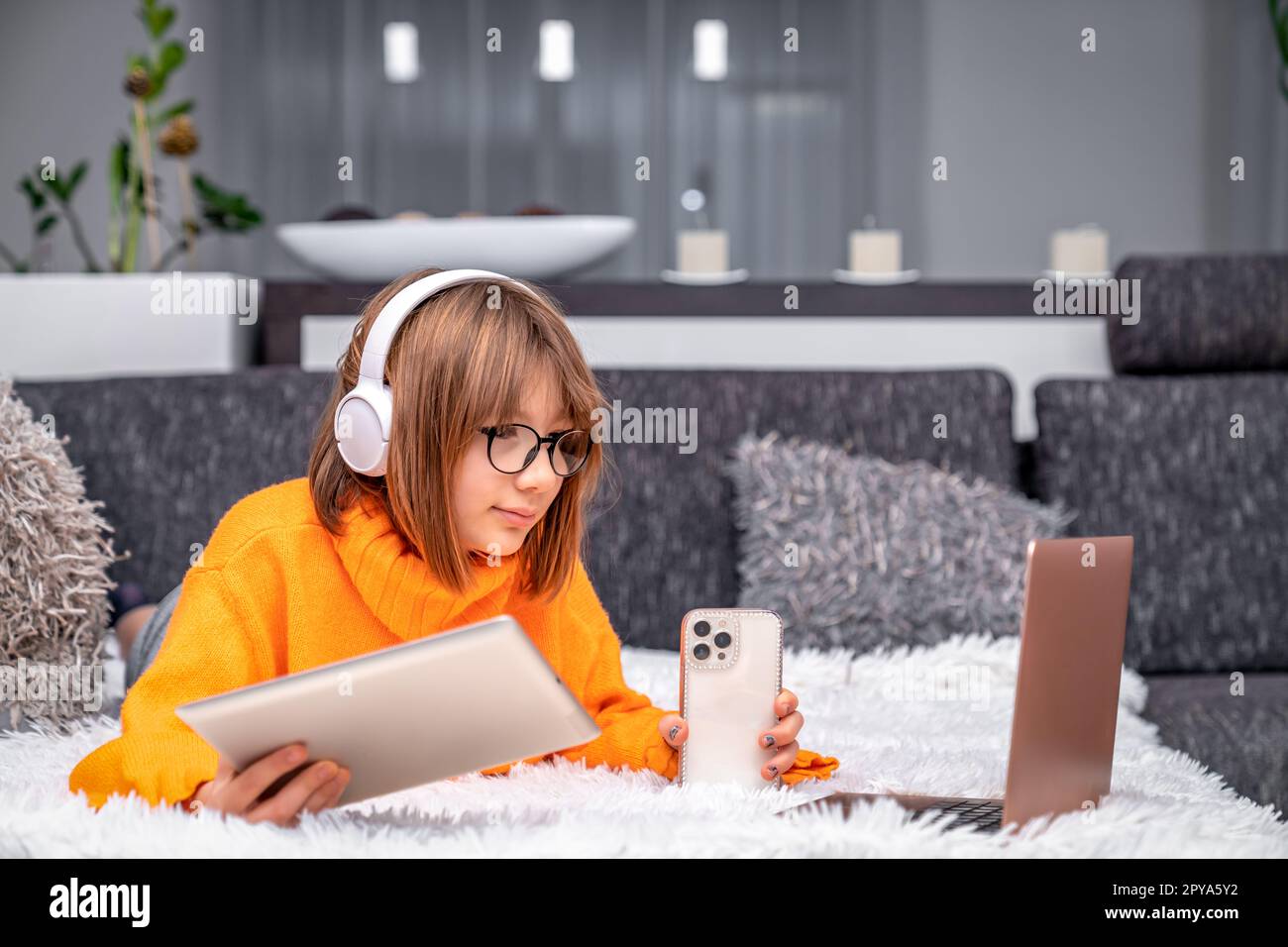 Moderne Technik und Kinder im Haushalt. Ein Mädchen mit kabellosen Kopfhörern kommuniziert online über Smartphone, Tablet und Laptop Stockfoto