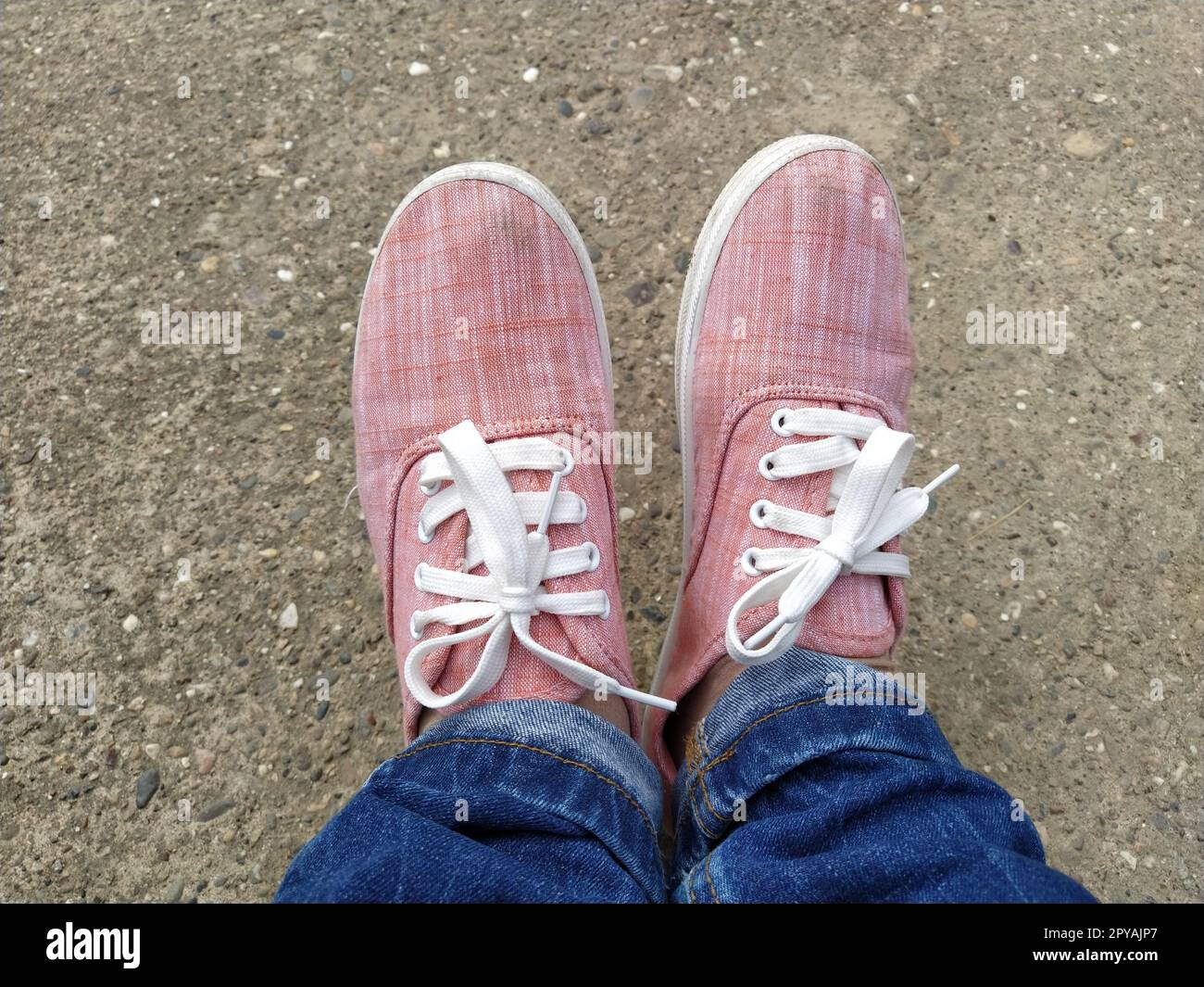 Pinkfarbene Sneaker mit weißen Schnürsenkeln an den Beinen. Blaue Jeans, darunter eingewickelt. Blick von oben. Sorgenfrei und sportlich. Stockfoto