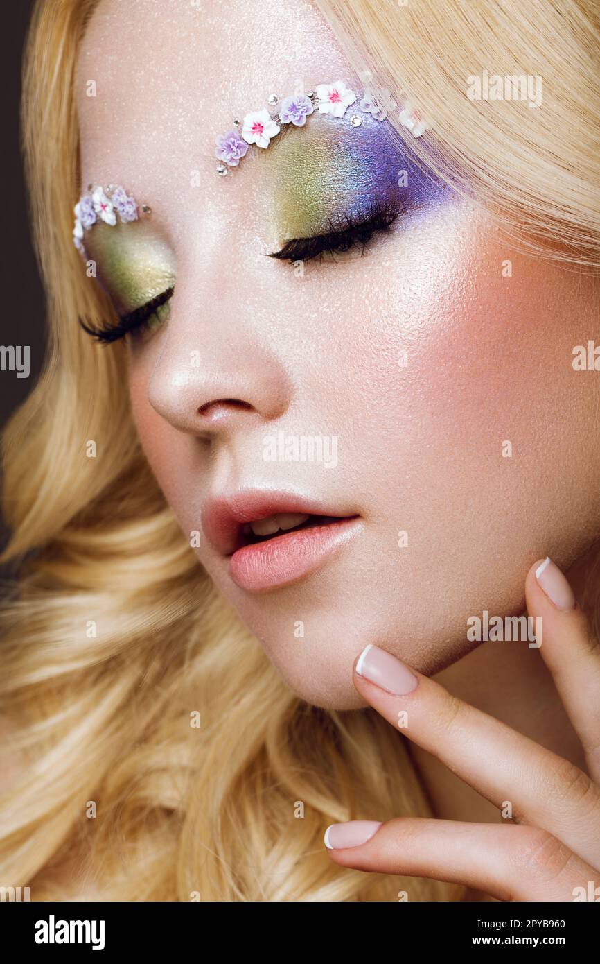Wunderschöne junge blonde Frau mit kreativer Make-up-Farbe, Locken und Blumen an den Augenbrauen. Schönheitsgesicht. Kunst-Make-up. Stockfoto