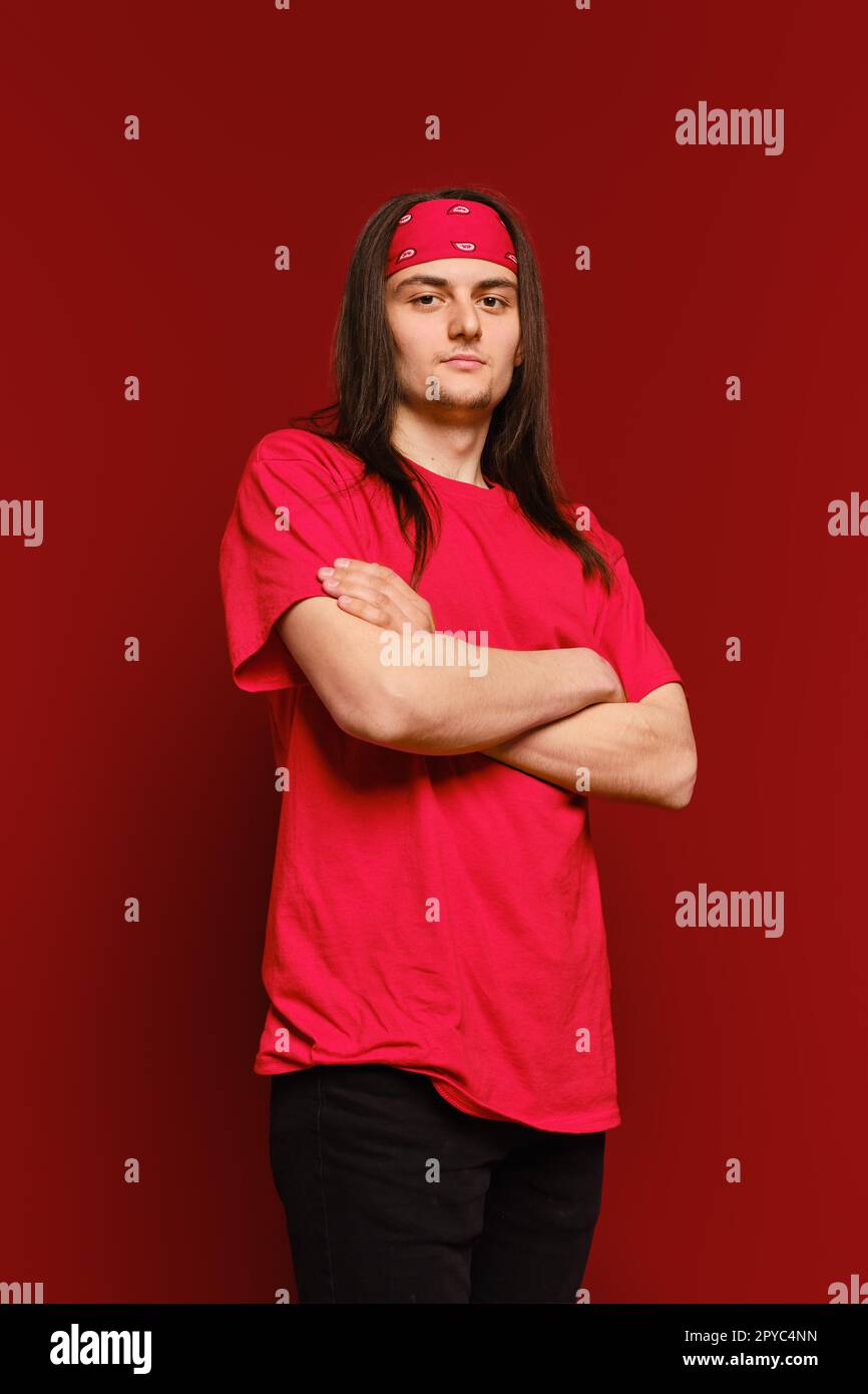 Ein junger erwachsener Mann steht in einem Studio mit verschränkten Armen und schaut selbstbewusst auf die Kamera vor rotem Hintergrund Stockfoto