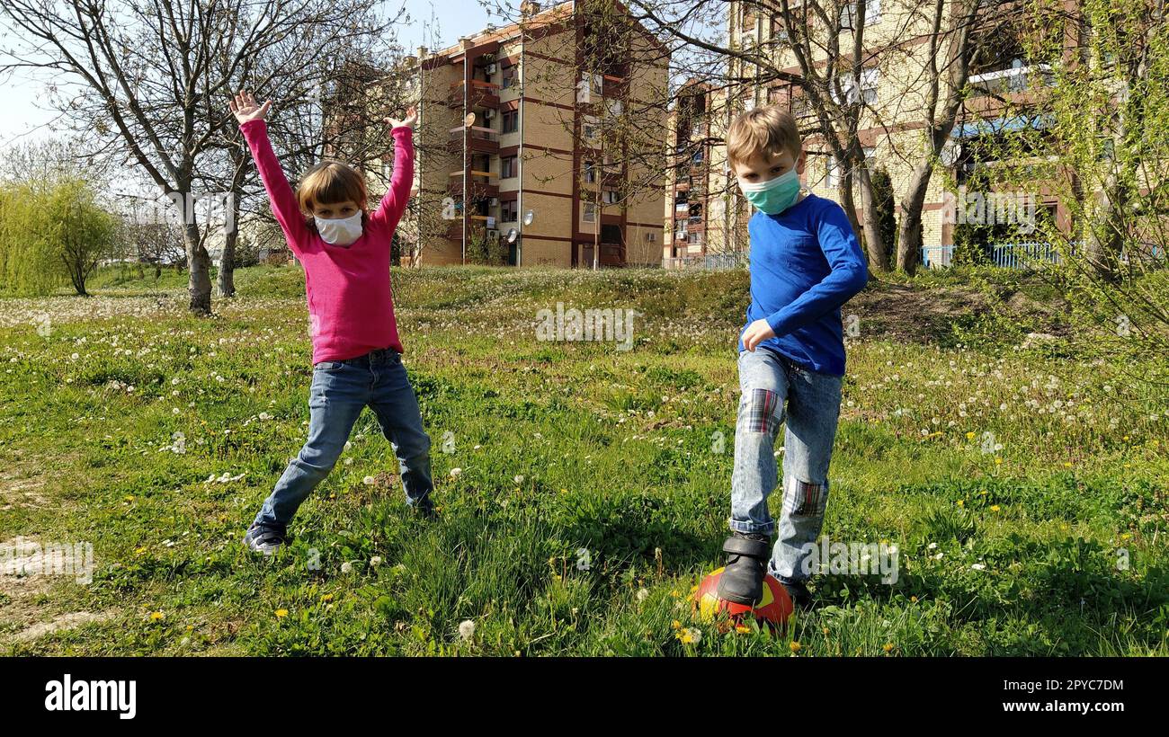 Ein Junge und ein Mädchen in schützenden chirurgischen Masken spielen Ball und tanzen auf dem grünen Gras. Schutz vor Luftkrankheiten. Die Wiederbelebung des Lebens und die Ankunft des Frühlings. Das Mädchen hob die Hände Stockfoto