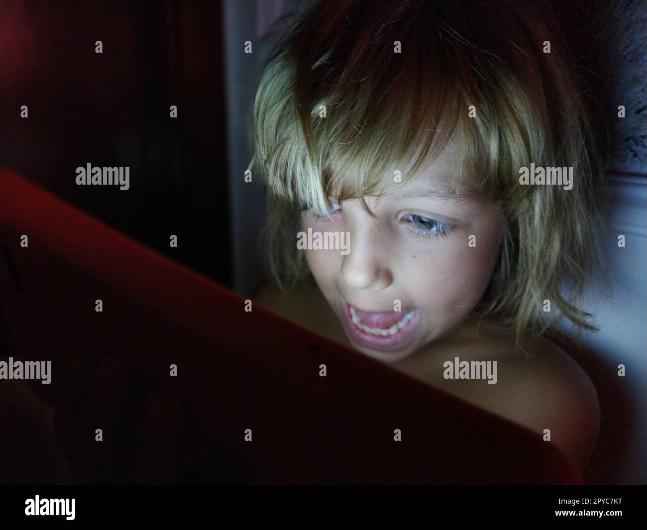 Süßes kleines Mädchen spielt mit einem Tablet-Computer. Ein 7-jähriges Kind mit lächelndem Gesicht und blondem Haar sieht in ein Gerät. Speicherplatz kopieren. Mädchen im Zimmer. Dunkle und helle Wand. Abends zu Hause Stockfoto