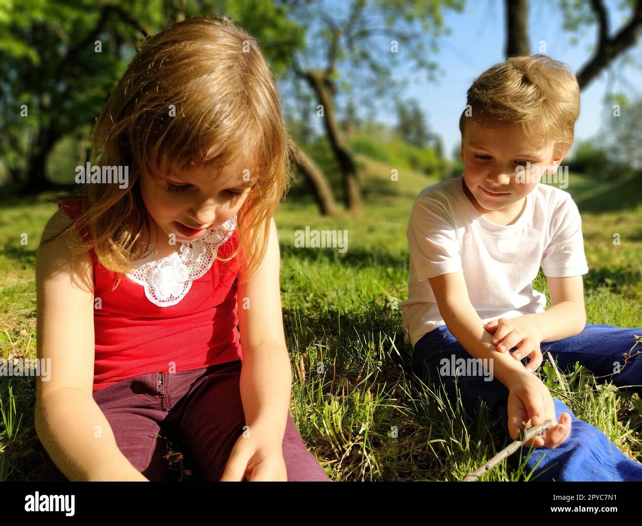 Zwei Kinder sitzen auf einer Wiese und langweilen sich vor Faulheit. Ein Mädchen im Alter von 6 Jahren und ein Junge im Alter von 7 Jahren spielen draußen in einem Stadtpark. Ein Junge in einem weißen T-Shirt, ein Mädchen in einem roten T-Shirt. Stockfoto