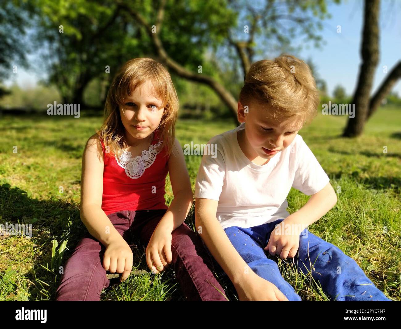 Zwei Kinder sitzen auf einer Wiese und langweilen sich vor Faulheit. Ein Mädchen im Alter von 6 Jahren und ein Junge im Alter von 7 Jahren spielen draußen in einem Stadtpark. Ein Junge in einem weißen T-Shirt, ein Mädchen in einem roten T-Shirt Stockfoto