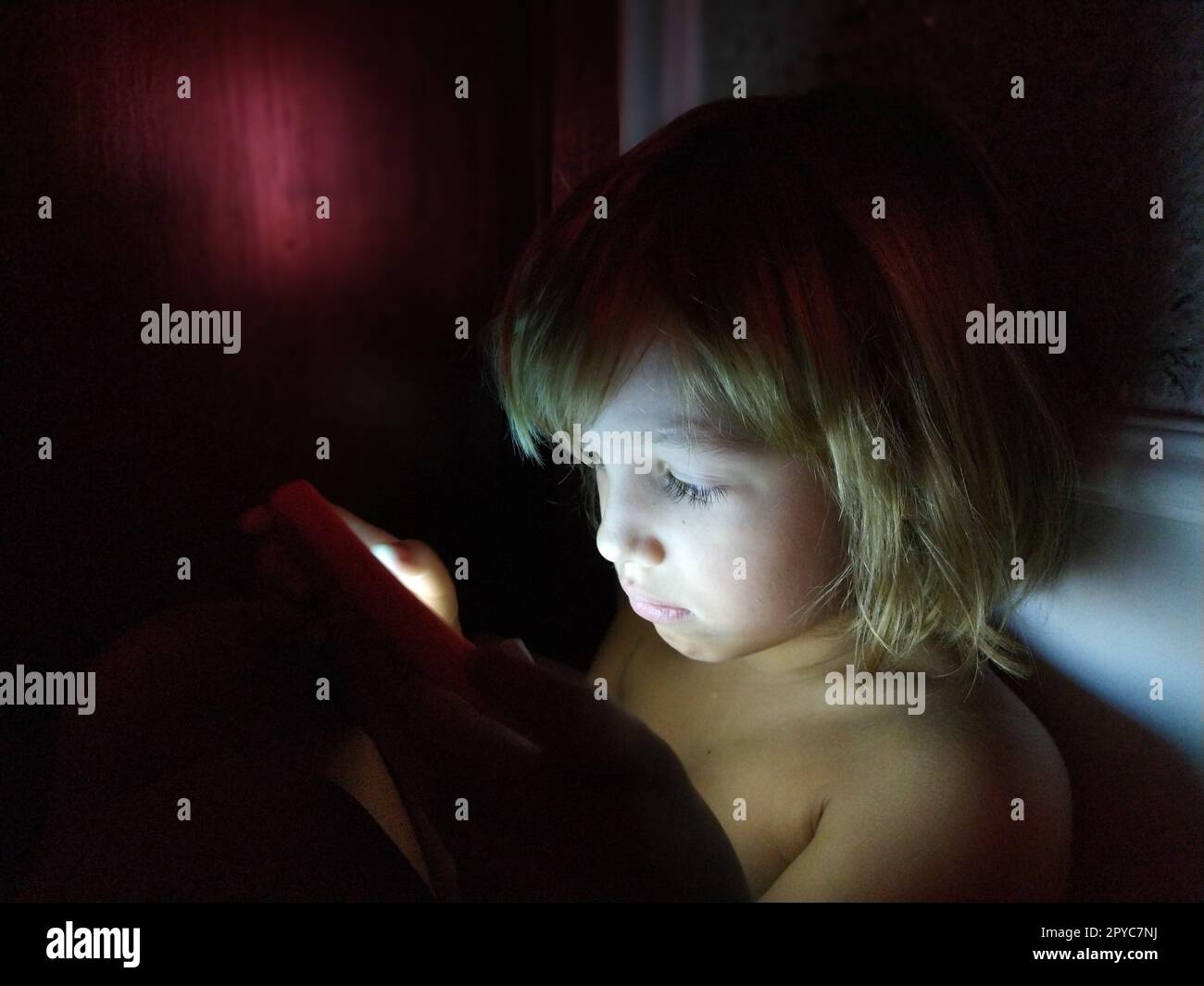 Süßes kleines Mädchen spielt mit einem Tablet-Computer. Ein 7-jähriges Kind mit einem ernsten Gesicht und blonden Haaren sieht in ein Gerät. Speicherplatz kopieren. Mädchen im Zimmer. Dunkle und helle Wand. Zu Hause in der Nacht Stockfoto