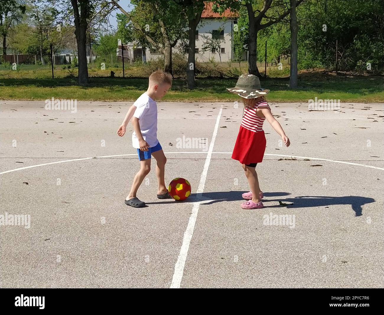 Kinder spielen draußen Ball. Ein Junge in einem weißen T-Shirt und ein Mädchen in einem roten Kleid spielen Ball auf einem Asphaltspielplatz. Mädchen mit Hut und breiter Krempe. Der Hof ist mit einem weißen Streifen bedeckt. Sommerzeit Stockfoto