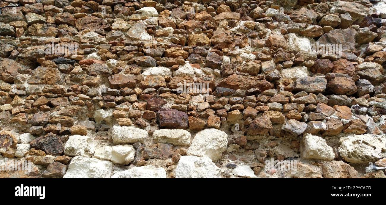 Steinmauer. Gestapelte große Ziegel und Kopfsteinpflaster in warmen Farben. Lose weiße Steine, die aus der Wandoberfläche hervorstehen. Stockfoto