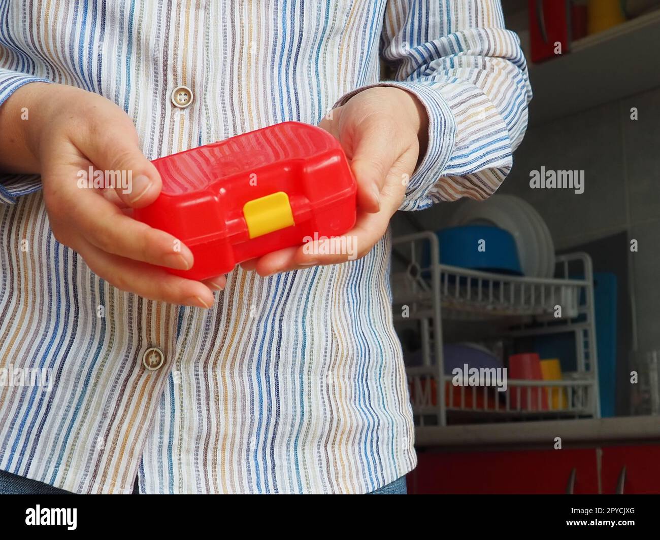 Weibliche Hände halten einen roten geschlossenen Plastikbehälter oder eine Verpackung für Schulessen. Küchenutensilien. Eine Frau in einem weißen gestreiften Hemd zeigt die Brotdose. Thema Lebensmittelhygiene und Recycling Stockfoto