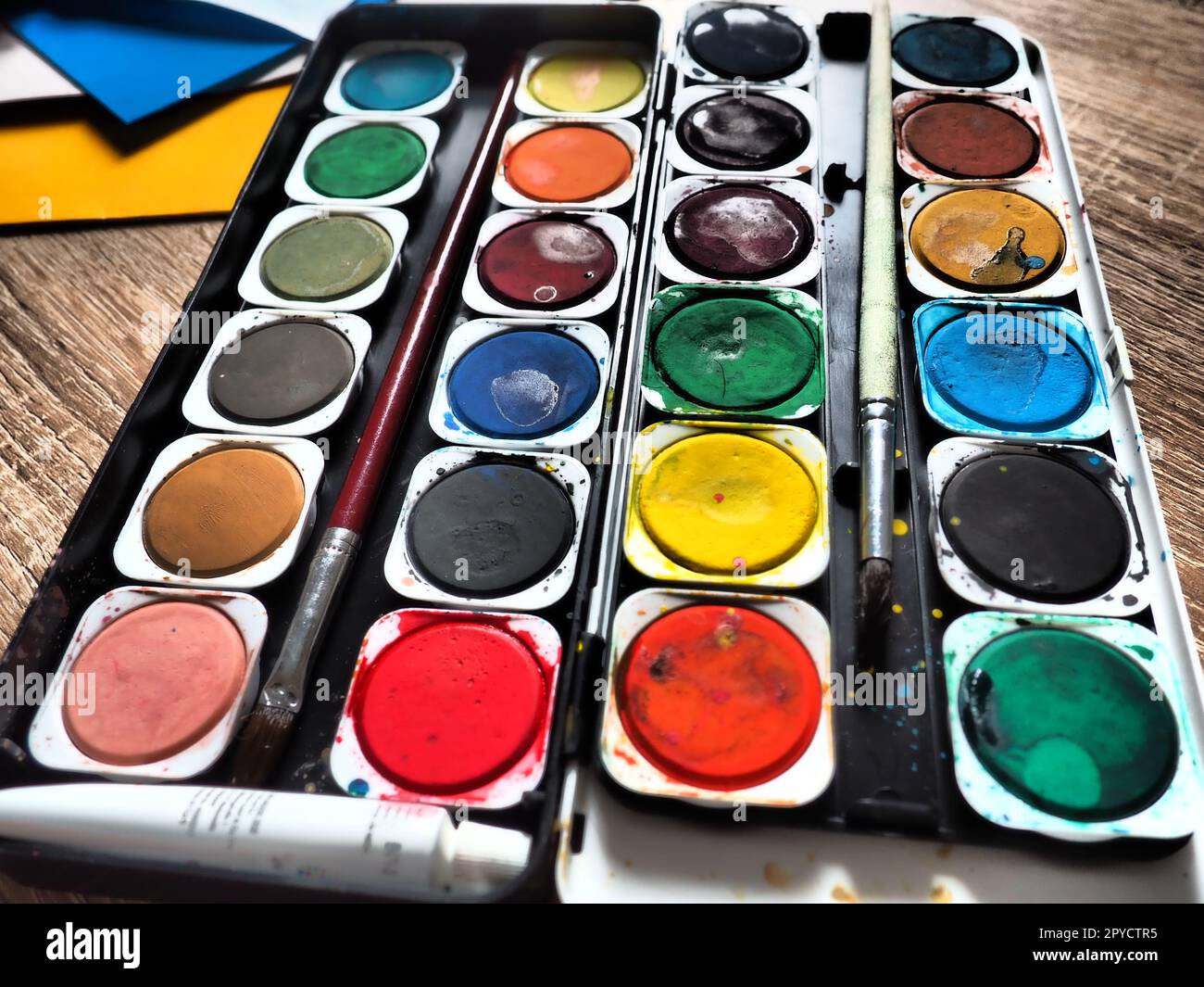 Ein Satz von Wasserfarben in verschiedenen Farben für das Zeichnen. Mehrfarbige runde Behälter mit heller Farbe. Werkzeuge für den Unterricht von Zeichnen und Arbeit. Farbiges Papier. Rot, Blau, Gelb, Grün. Stockfoto