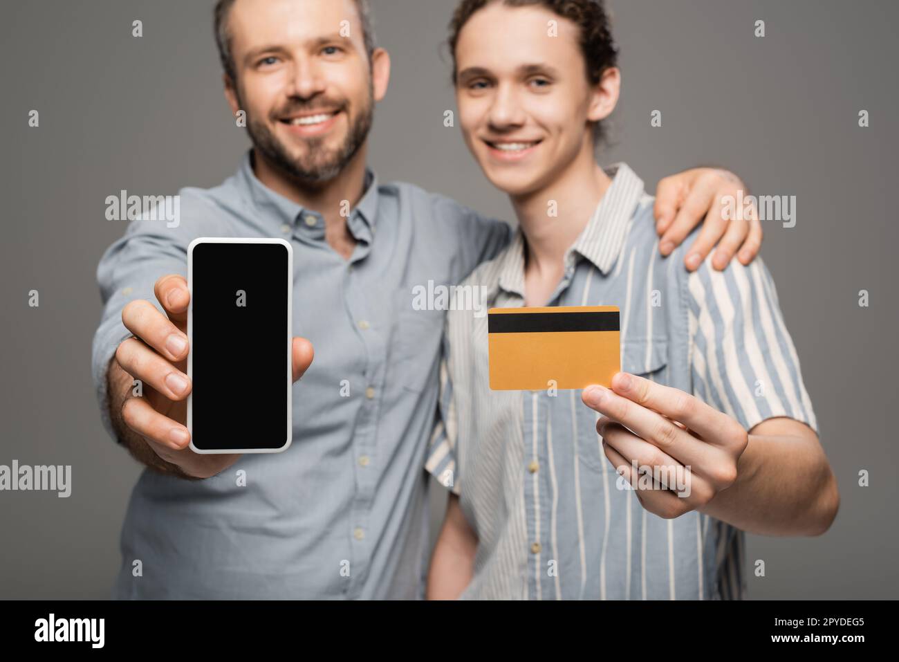 Überglücklich, der Vater hielt ein Smartphone mit leerem Bildschirm neben einem glücklichen Teenager-Sohn mit Kreditkarte in der Hand isoliert auf grauem Stockbild Stockfoto