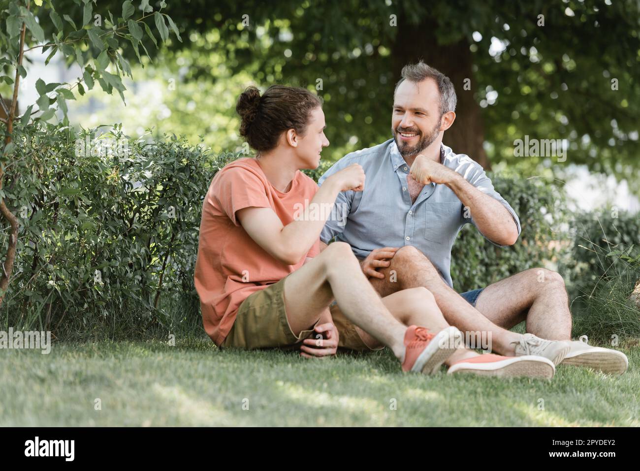 Glücklicher Vater und Sohn beim Fauststoß, während sie auf dem grünen Rasen im Park saßen, Stockbild Stockfoto
