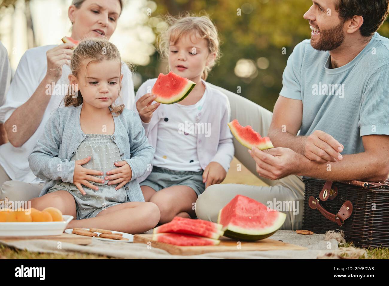 Park, Picknick und Paar mit Kindern und Obst auf der Decke im Garten für einen schönen Sommer mit der Familie. Natur, Liebe und Entspannung Mittagessen auf dem Rasen mit Eltern und Kindern mit Lächeln im Urlaub. Stockfoto