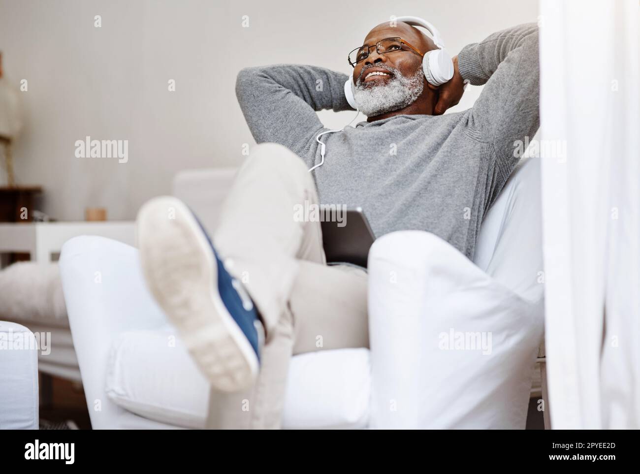 Dieses Lied erinnert mich an meine goldenen Tage. Ein gutaussehender Senior-Mann, der Musik auf einem Tablet hört, während er sich zu Hause auf dem Sofa entspannt. Stockfoto