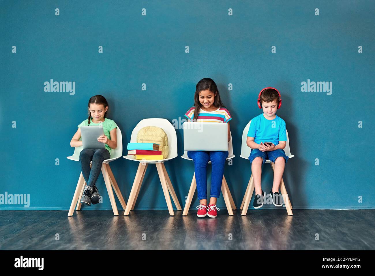 Mit drahtlosen Geräten aufwachsen. Studioaufnahme von Kindern, die auf Stühlen sitzen und drahtlose Technologie verwenden, vor blauem Hintergrund. Stockfoto