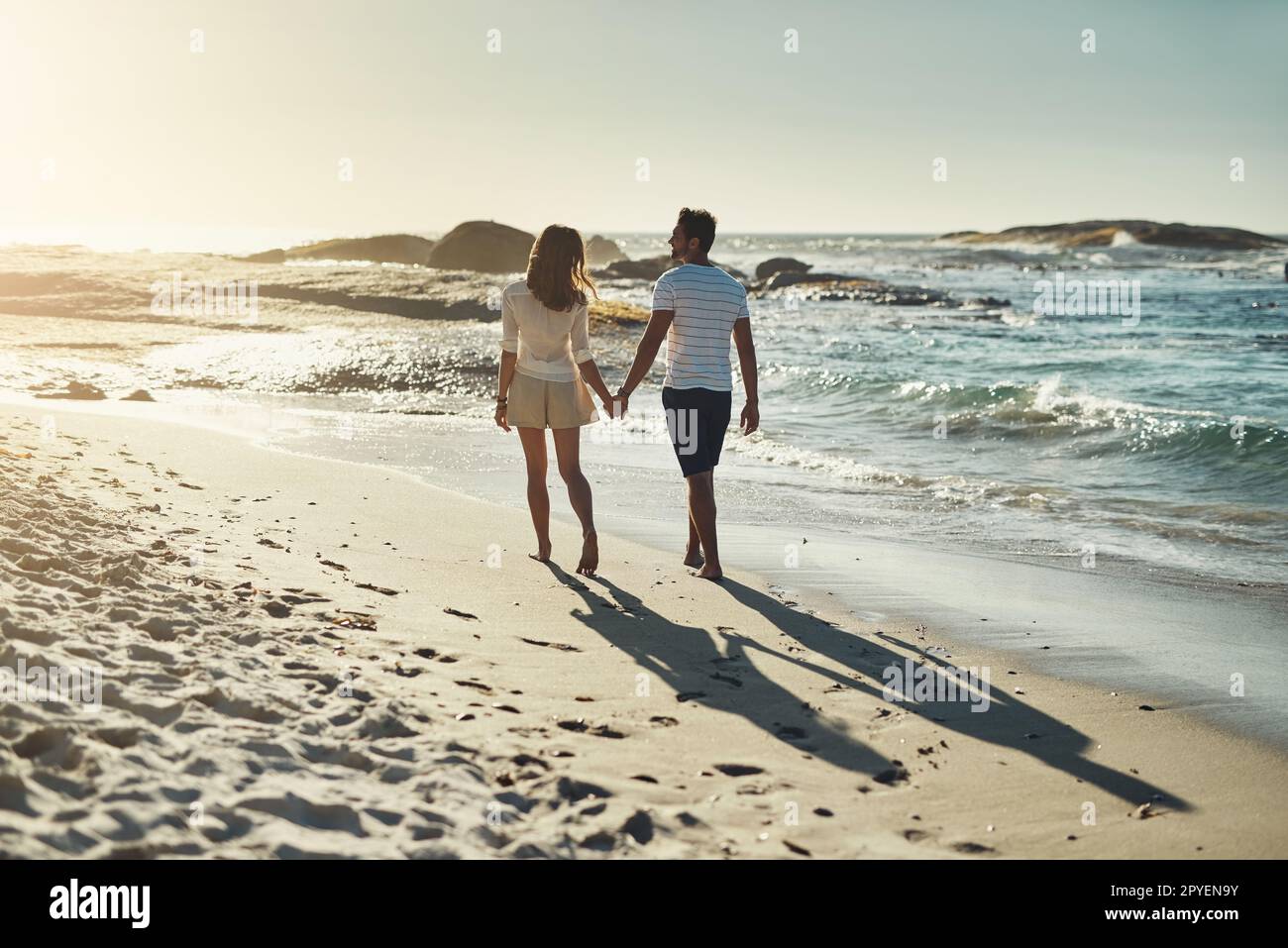 Sie genießen lange Spaziergänge am Strand. Rückblick auf ein sorgenfreies junges Paar, das Hand in Hand am Strand entlang geht. Stockfoto