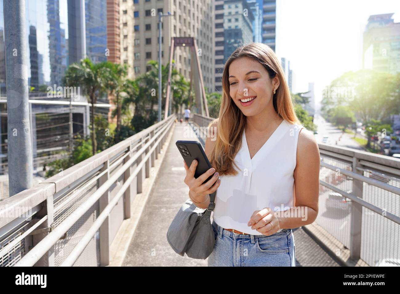 Porträt eines lächelnden Mädchens auf der Fußgängerbrücke, das sein Smartphone in der modernen, nachhaltigen Metropole Sao Paulo, Brasilien, sieht Stockfoto