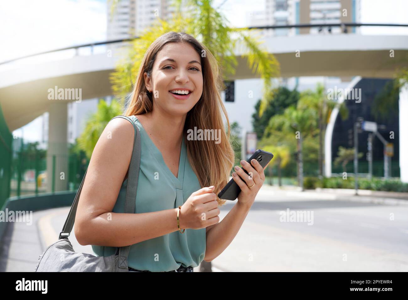 Halblanges Porträt einer fröhlichen brasilianischen Frau auf der Straße mit Smartphone. Ein wunderschönes Millennials-Mädchen, das in der Stadt in der Hand eines Mobiltelefons in die Kamera schaut. Stockfoto