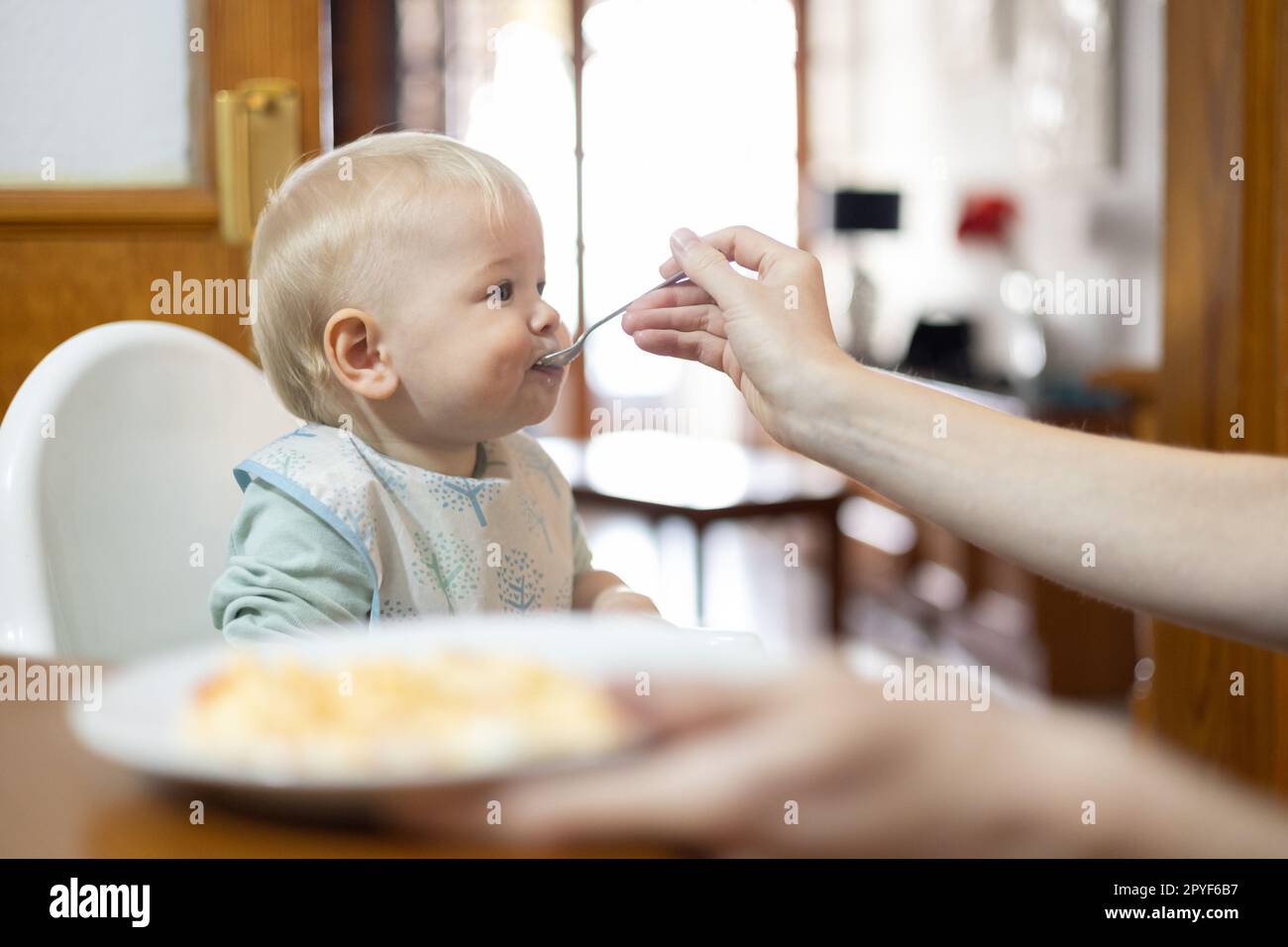 Mutterlöffel füttert ihr Baby Kind, das in einem Hochstuhl am Esstisch in der Küche zu Hause sitzt Stockfoto