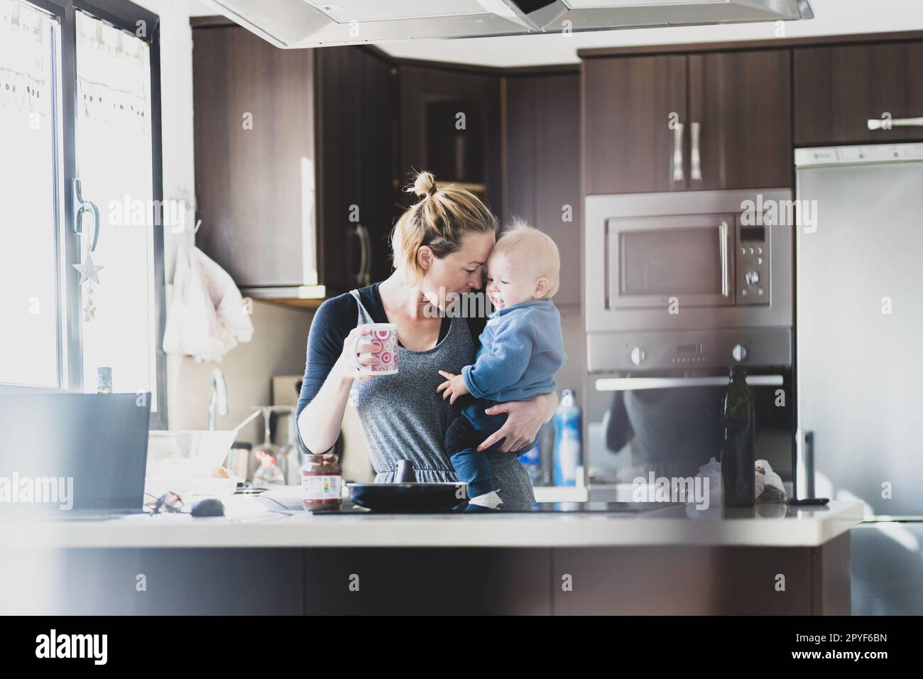 Glückliche Mutter hält ihren kleinen kleinen kleinen Jungen, während sie morgens Kaffee trinkt und Pfannkuchen zum Frühstück in der Hausküche macht. Familienleben, häusliches Lebenskonzept. Stockfoto
