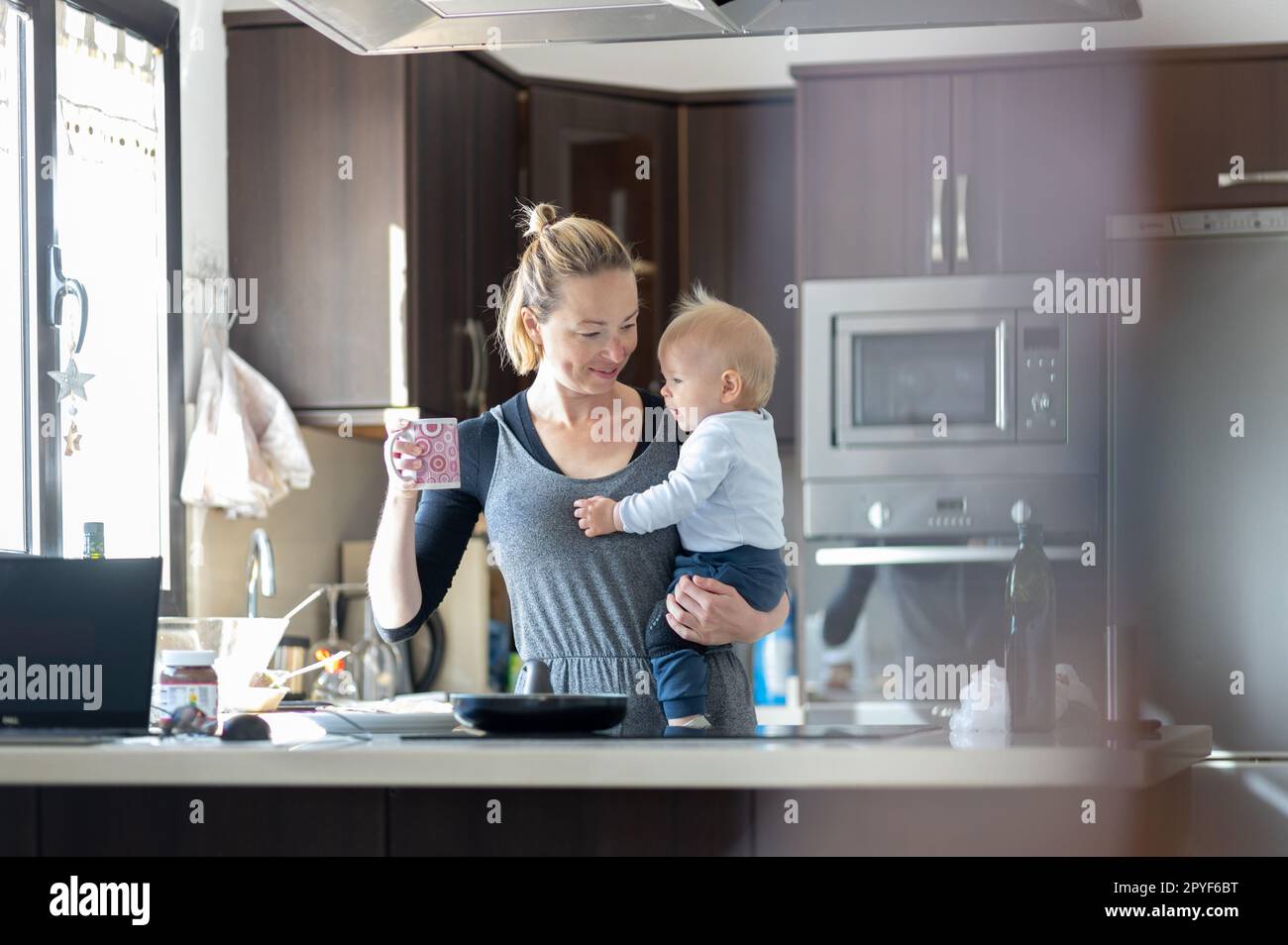 Glückliche Mutter hält ihren kleinen kleinen kleinen Jungen, während sie morgens Kaffee trinkt und Pfannkuchen zum Frühstück in der Hausküche macht. Familienleben, häusliches Lebenskonzept. Stockfoto