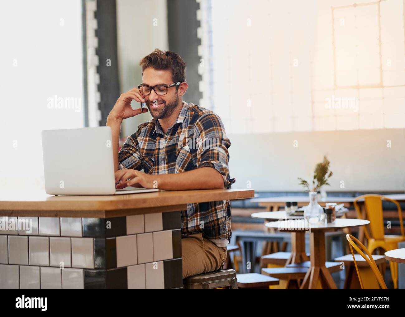 Ich arbeite heute von meinem mobilen Büro aus. Ein glücklicher junger Unternehmer geht an sein Handy, während er in einem Café an seinem Laptop arbeitet. Stockfoto