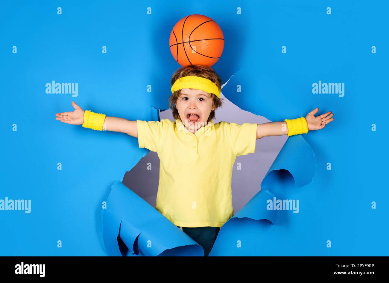 Basketballspiel. Überraschtes Kind mit Basketballball auf dem Kopf. Kleiner Basketballer in Sportbekleidung, der durch Papier schaut. Aktiver Sport. Der Junge spielt Stockfoto