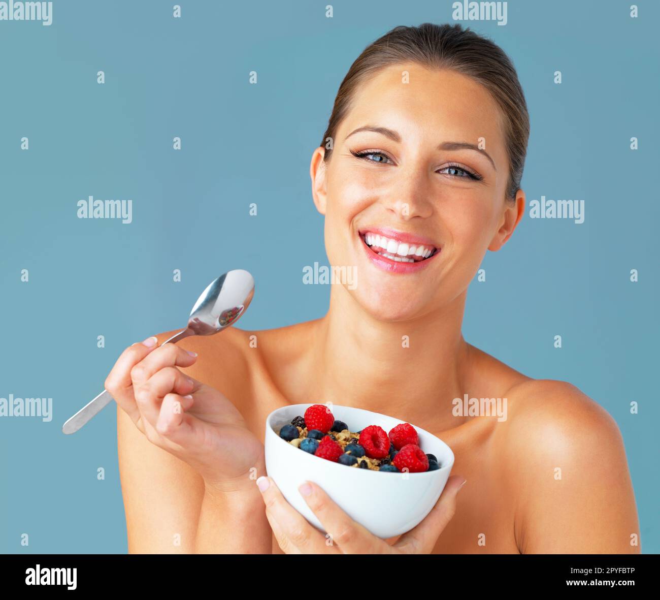 Essen Sie gesund, seien Sie glücklich. Studioaufnahme einer attraktiven jungen Frau, die eine Schüssel Müsli und Obst isst, vor blauem Hintergrund. Stockfoto