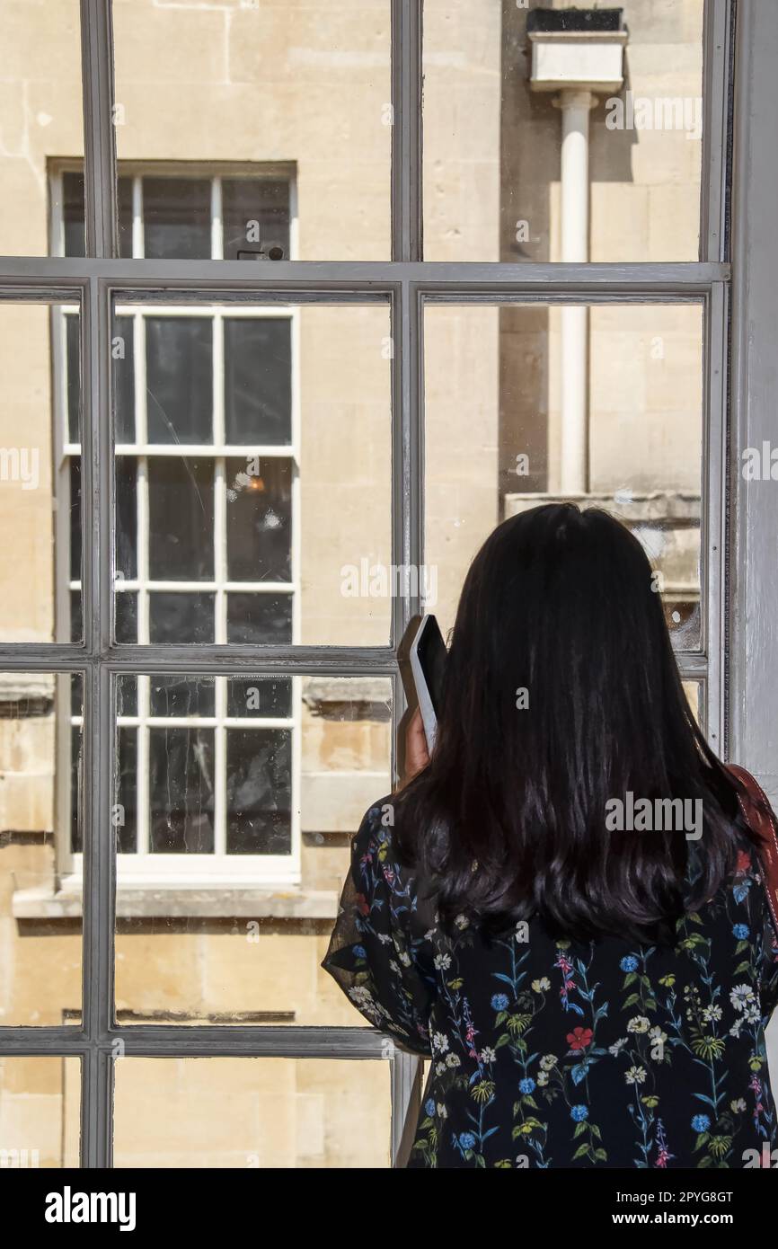 Rückansicht - Mädchen mit hübschem Blumenkleid und langen schwarzen Haaren spricht am Handy, während sie aus dem Holzfenster auf das alte Gebäude gegenüber blickt - selig Stockfoto