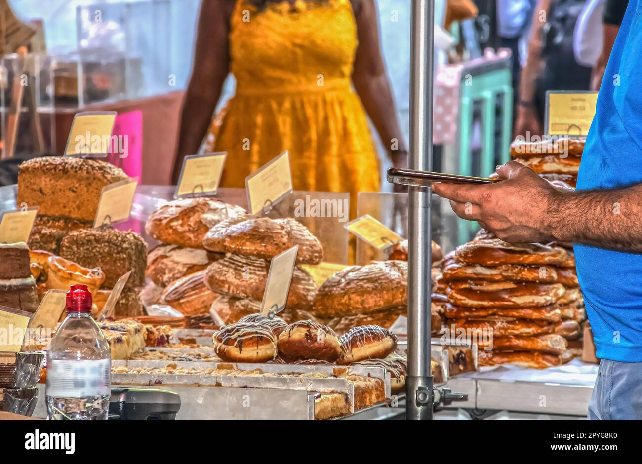 Bäckerei auf dem Markt mit Schwerpunkt auf Vordergrund - Verkäufer überprüft Telefon, wenn Frau in gelbem Kleid nähert - verkürzt und nicht wiederzuerkennen Stockfoto