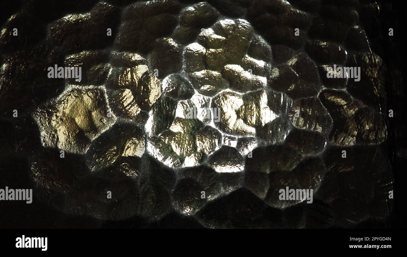 Wellglas, Rücklichtquelle. Spiel von Blendungen und Reflexionen auf einer transparenten, unebenen Oberfläche. Dunkle und helle abstrakte asymmetrische, mehrfach beleuchtete Ebenen. Abstrakter, holpriger goldener Hintergrund. Stockfoto