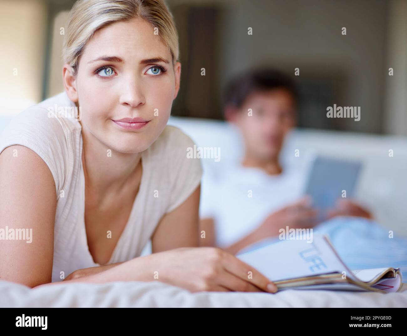 Ich fühle es nicht. Eine gelangweilte junge Frau, die im Bett liegt und eine Zeitschrift liest, während ihr Mann ein digitales Tablet im Hintergrund benutzt. Stockfoto