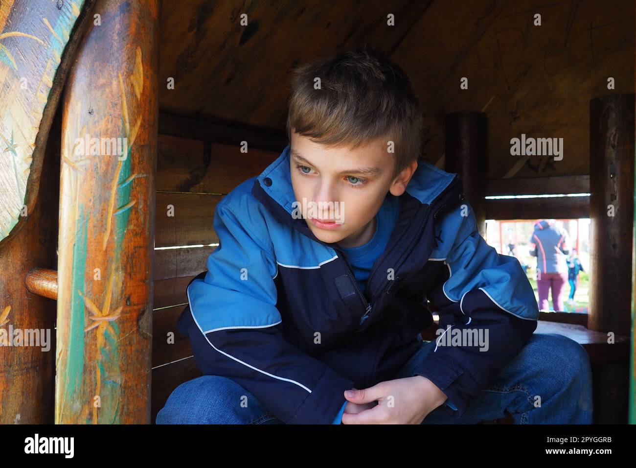 Ein 10-jähriger Kaukasier mit blonden Haaren und grauen Augen schaut aus der Tür eines Holzhauses auf einem Kinderspielplatz. Peekaboo. Das Kind versteckte sich. Hübsches Gesicht. Der Junge sitzt in einem kleinen Haus. Stockfoto