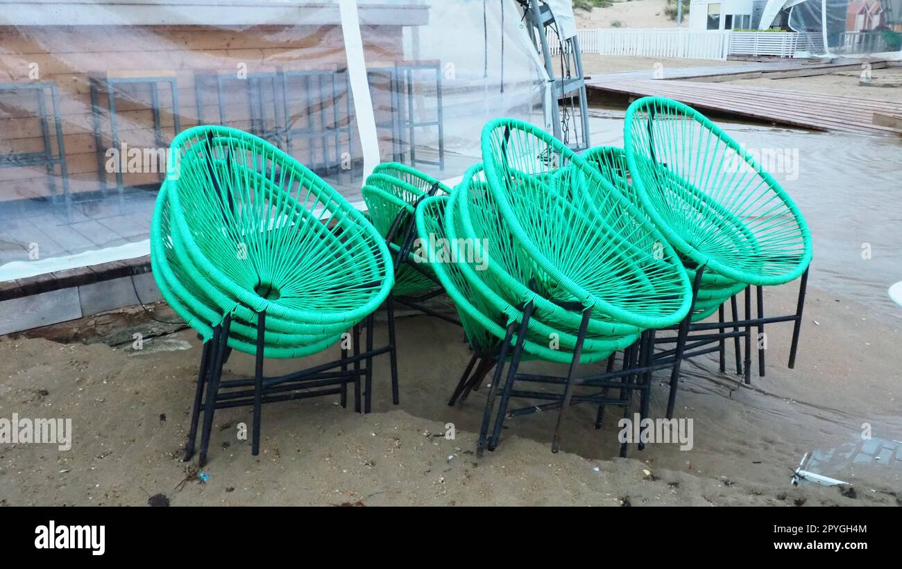 Korbgrüne Plastikstühle mit schwarzen Beinen in einem Café an einem Sandstrand nach einem Sturm. Die Möbel sind übereinander gestapelt, da das Strandcafe geschlossen ist. Ende der Strandsaison im Hotel Stockfoto