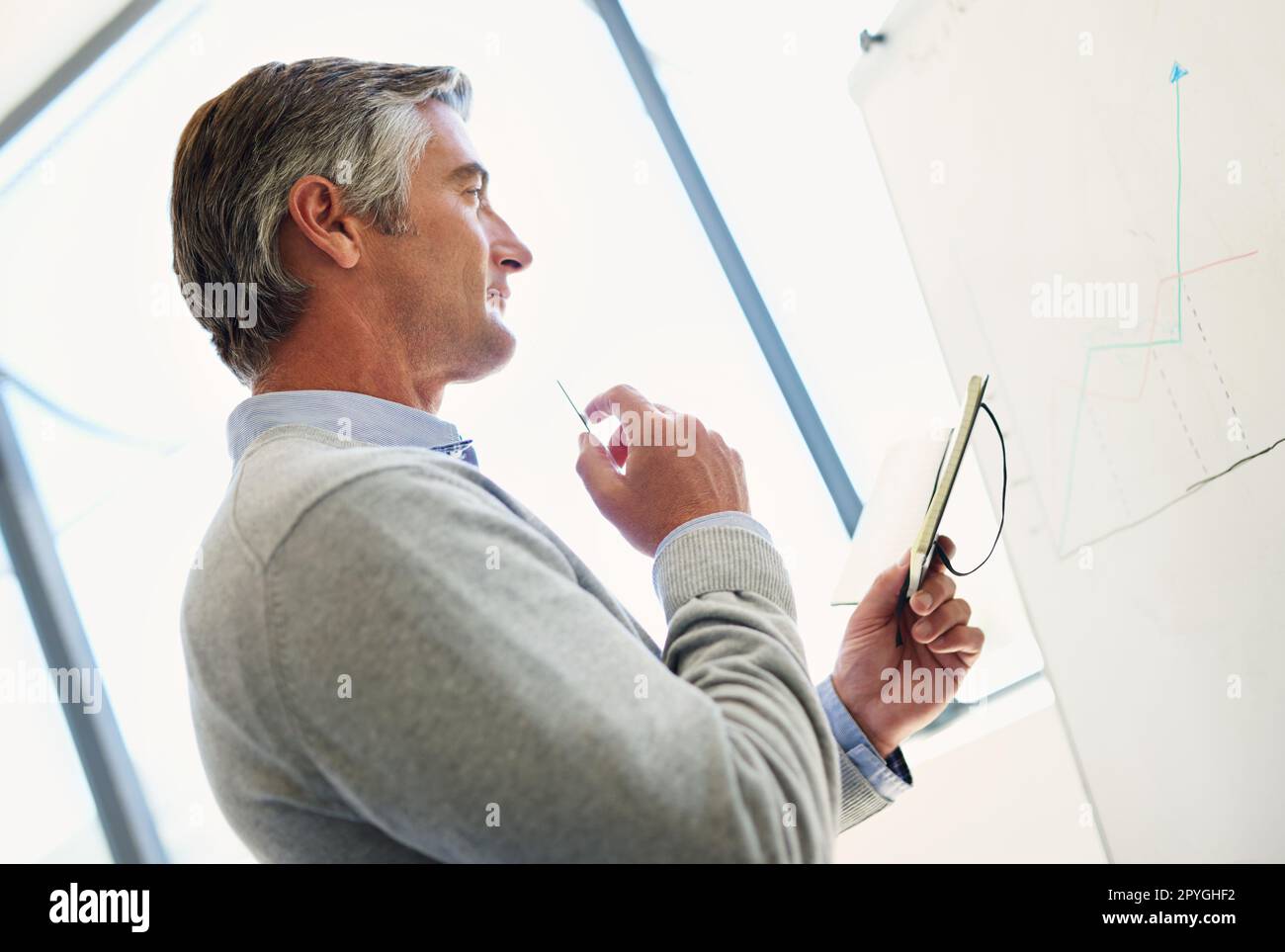 Ich sehe mir die Zahlen an. Ein reifes Unternehmen, das aufmerksam aussieht, während es an einem Whiteboard mit einem Notebook steht. Stockfoto