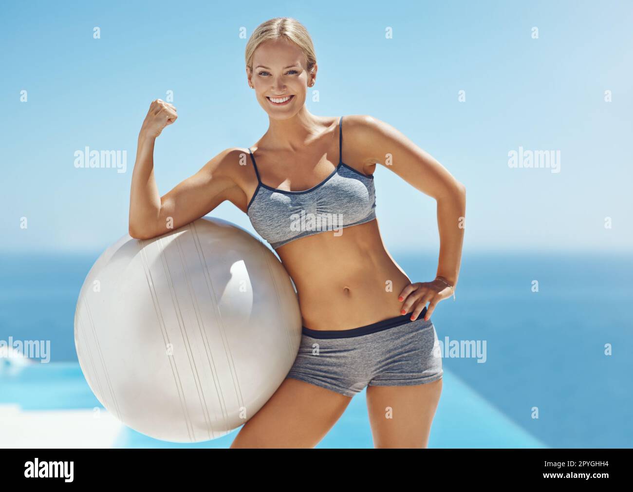 Ich bin abs-stehend. Eine attraktive junge Frau, die nach ihrem Workout ihren Bizeps auf einem Sportball am Pool bewegte. Stockfoto