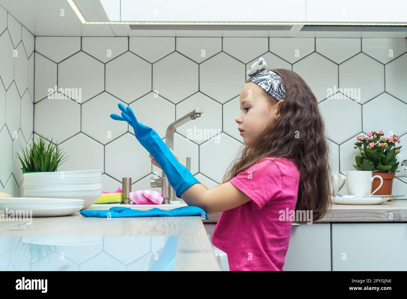Nettes kleines Mädchen, das blaue Haushaltshandschuhe anzieht, um Geschirr abzuspülen. Hausreinigung, persönliche Schutzausrüstung. Stockfoto
