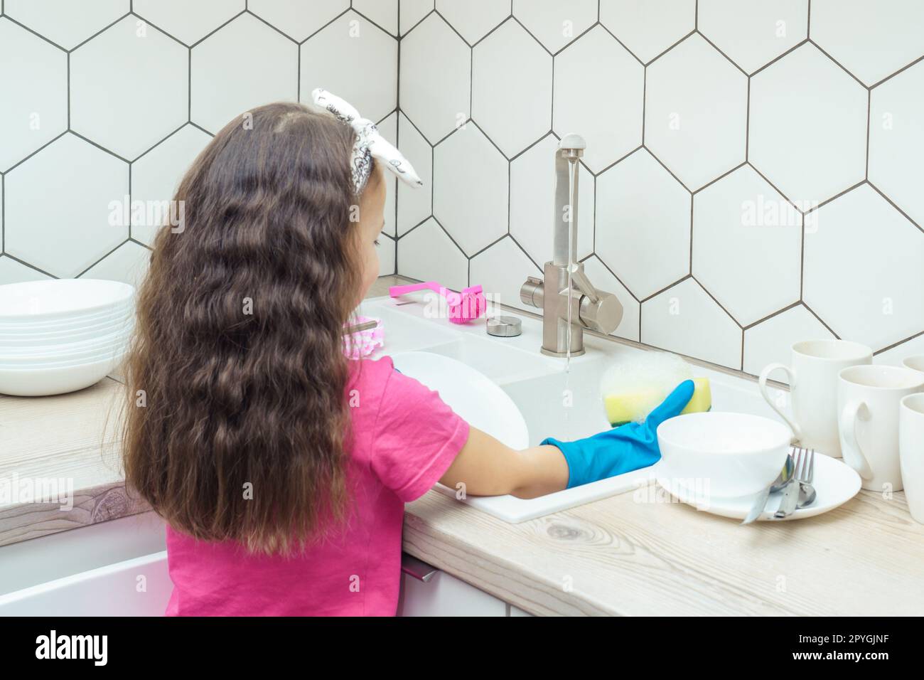 Ein kleines Mädchen in Haushaltshandschuhen, das Geschirr mit einem schaumigen Spülschwamm im Spülbecken unter Wasserstrahl abspült. Stockfoto