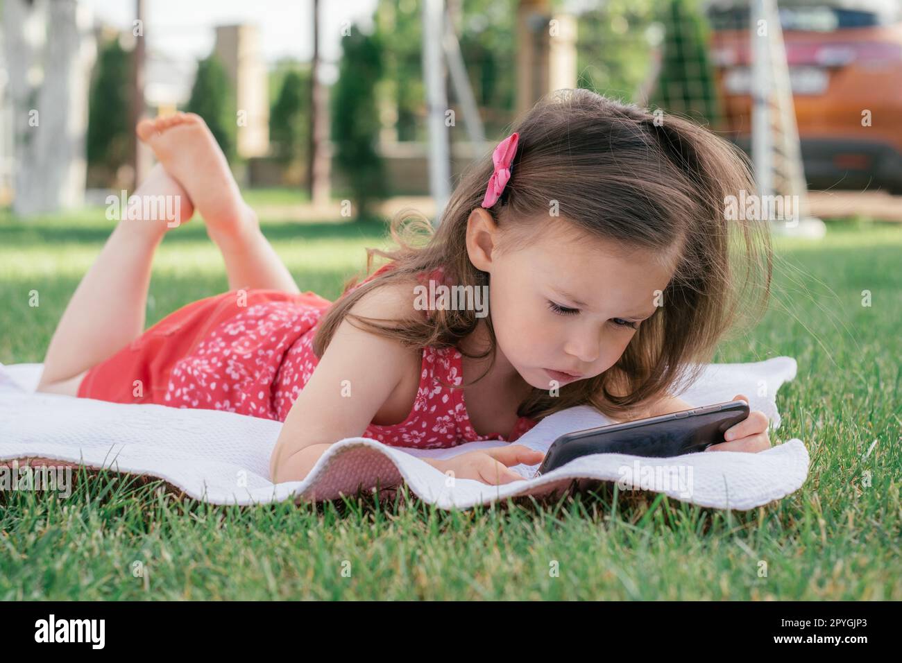Das kleine 3-4-Mädchen in roten Kleidern liegt auf einer Decke auf grünem Gras und schaut in das Handy. Kinder, die Gadgets benutzen Stockfoto
