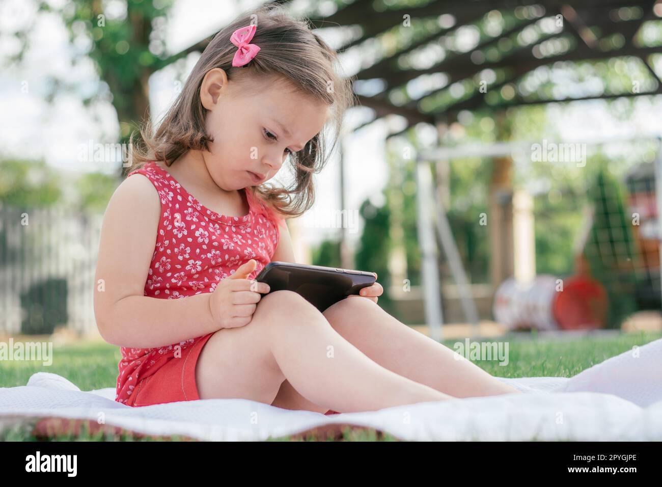 Das kleine 3-4-Mädchen in roten Kleidern sitzt auf einer Decke auf grünem Gras und schaut in das Handy. Kinder, die Gadgets benutzen Stockfoto