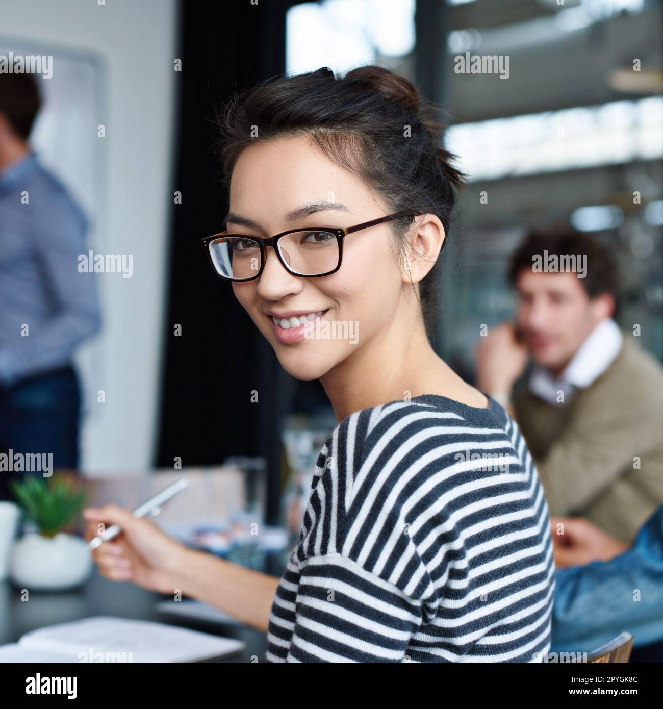 Die perfekte Arbeitsumgebung. Porträt einer attraktiven jungen Frau, die im Büro mit Kollegen im Hintergrund sitzt. Stockfoto