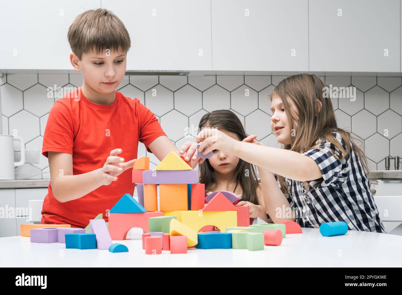 Positive, fokussierte Kinder von Jungen und zwei Mädchen, die mit bunten Bausteinen spielen und eine Burg aus geometrischen Figuren bauen Stockfoto
