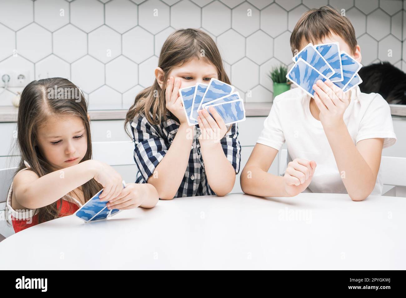 Lustiges Foto, drei Kinder, die sich blaue Spielkarten in den Händen ansehen, mit Enttäuschung und Sorge, sitzen am weißen Tisch Stockfoto