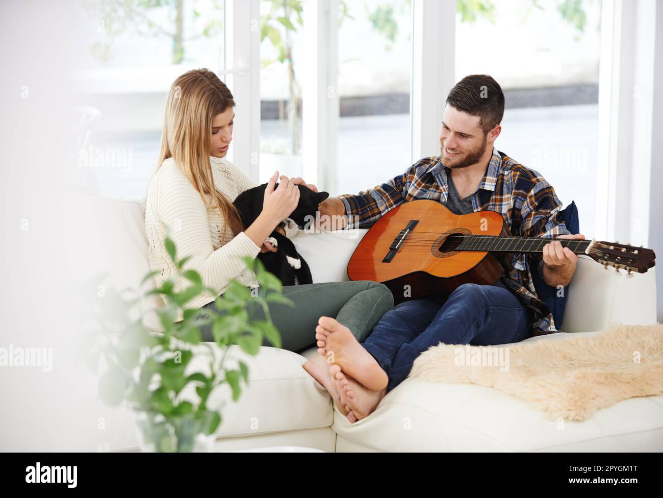 Die entspannendsten Nachmittage. Ein junger Mann, der für seine Freundin Gitarre spielt, während er auf dem Sofa sitzt. Stockfoto