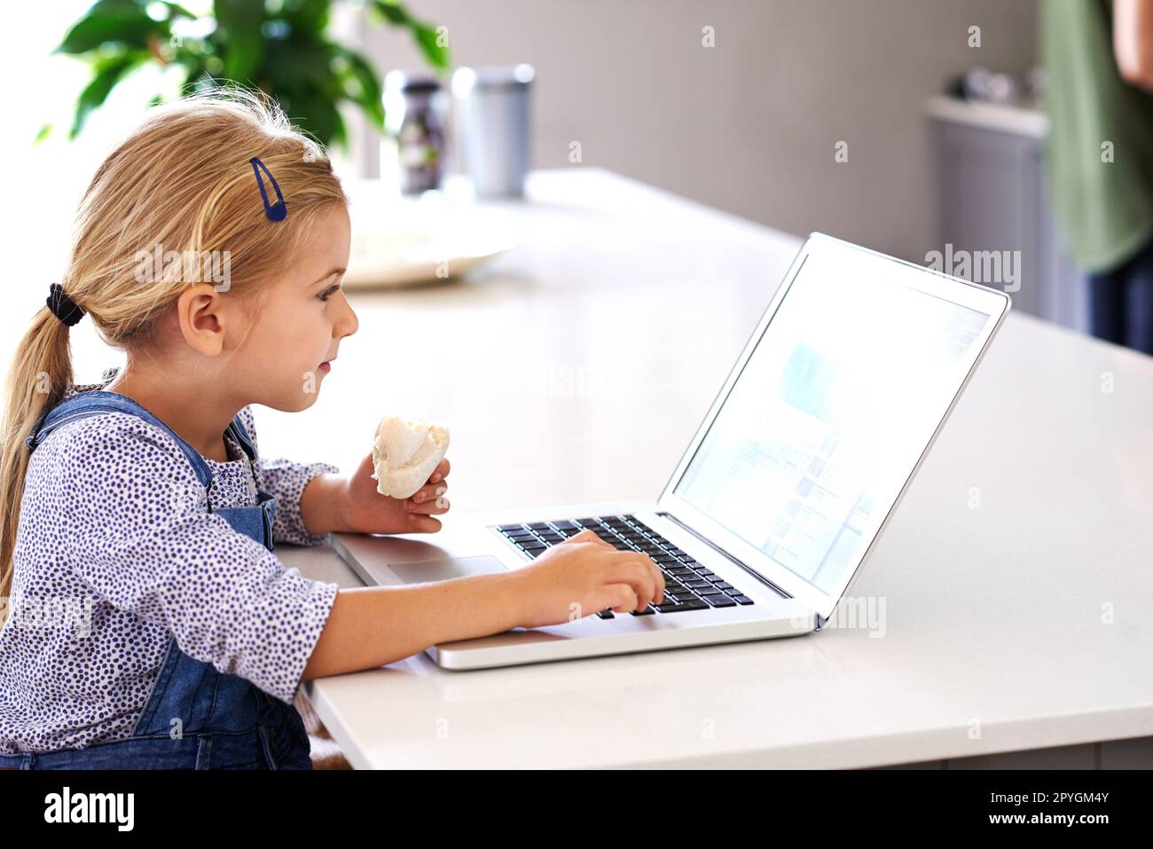 Es kann schwierig sein, das Mittagessen durchzuarbeiten. Ein süßes kleines Mädchen, das einen Laptop benutzt, während es einen Snack isst. Stockfoto